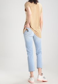 Camicia beige chiaro a maniche corte abbinata a jeans a vita alta azzurri con ricamo di arcobaleno e stelle sulla tasca posteriore, indossata con sneakers bianche.