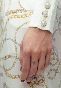 Material alb cu model de lanțuri aurii și gri. Mâneci cu mansetă, având trei nasturi albi cu accente aurii. Mână tatuată. Oja roșie pe unghii.