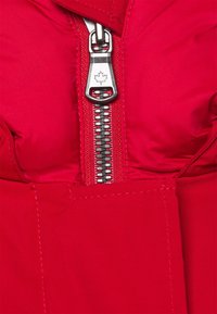 Rode ritsjack met een glanzende zilveren rits, een esdoornbladlogo, een geborduurde textuur en versterkte stiksels op de stof.