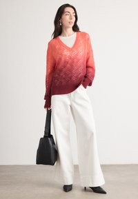 Pull en tricot dégradé rouge à orange avec motif en zigzag, associé à un pantalon large blanc et un sac à main noir. Des talons complètent l'ensemble.