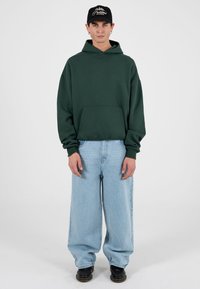 Roheline kapuutsiga dressipluus esistaskuga, kombineeritud helesiniste laiade pükstega. Modell kannab musta nokamütsi valge tekstiga. Mustad jalanõud.