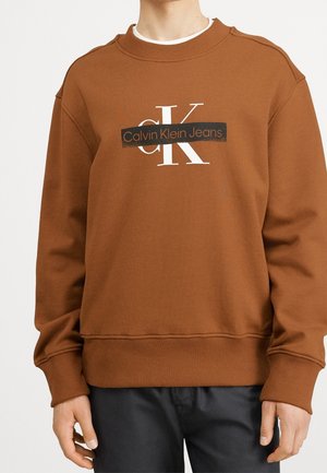 Persona con sudadera de color marrón oxidado con el logotipo "CK Calvin Klein Jeans" en blanco y negro en el pecho, combinada con pantalones negros.