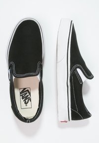 Svarta slip-on kanvas skor med vita sömmar och gummisulor, med en klassisk design och Vans-logotyp på innersulan.