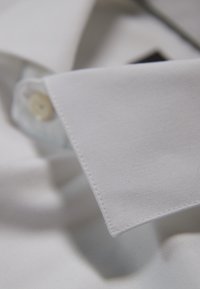 Primo piano del colletto di una camicia di cotone bianca, caratterizzato da una texture liscia, un singolo bottone e cuciture precise lungo i bordi.