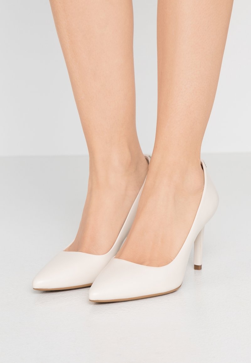 Paire de jambes portant des chaussures à talons hauts blanches à bout pointu sur un sol gris clair avec un fond gris clair uni.