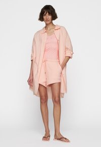 Camicia button-up leggera color pesca con strisce bianche, indossata sopra un top coordinato e pantaloni corti. Presenta una vestibilità comoda e una vita con lacci.