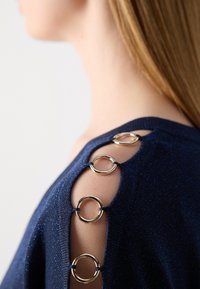 Navyblaues Strickoberteil mit runden goldfarbenen Hardware-Details an den Schultern und ausgeschnittenen Elementen, die Textur und Design hervorheben.