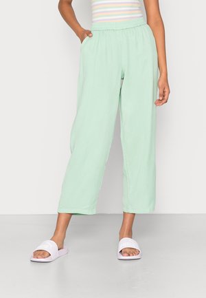 Calças largas, leves, de cor verde menta, com uma cintura elástica e bolsos laterais, combinadas com sandálias brancas sem traseira.