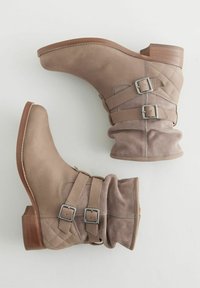 Botas de tobillo beige con parte superior de cuero, acentos acolchados, dos correas ajustables con hebillas y un tacón de bloque de madera. Superficies suaves y texturizadas.