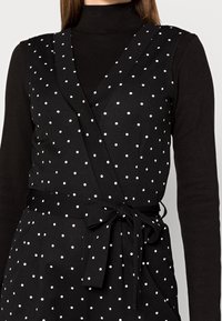 Svart wrap-top med vita prickar, med en djup V-ringning, ärmlös design och ett knytbälte i midjan. Slät textur.