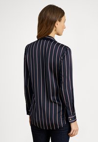 Sieviete ar brūniem matiem, valkā tumši zilu garroku krekla ar baltām un sarkanām vertikālām svītrām, skatās prom pret vienkrāsainu fonu.