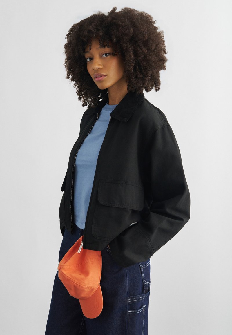 Young woman with curly hair wearing a black jacket over a blue shirt, dark pants, holding an orange cap against a plain background.