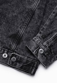 Gros plan du poignet et de la ceinture d'une veste en denim noire avec des boutons métalliques étiquetés « DENIM » et des détails de coutures visibles.