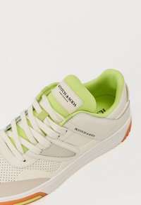 Weiße und hellgrüne Sneaker mit einer strukturierten Oberseite, durchbrochenen Details, cremefarbenen Schnürsenkeln und orangefarbenen Akzenten an der Sohle. Markenetikett sichtbar.