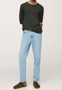 Personne portant une chemise à manches longues vert foncé, un jean bleu clair et des chaussures marron sans lacets, debout avec les mains derrière le dos.