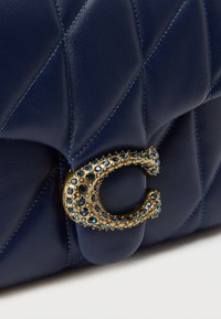 Τσάντα χειρός από δέρμα στο χρώμα του ναυτικού, με ανάγλυφο μοτίβο, που διαθέτει κούμπωμα σε χρυσό τόνο διακοσμημένο με μπλε πολύτιμους λίθους και υφασμένα στοιχεία.