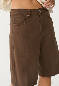 Mujer vestida con pantalones cortos de mezclilla marrón de tiro alto con bolsillos frontales y una blusa corta de encaje blanca, con la mano descansando cerca de la cintura.