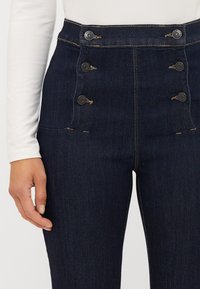 Jeans de cintura alta azul-escuro com uma textura de denim suave, apresentando seis botões decorativos e detalhes de costura em contraste subtil.