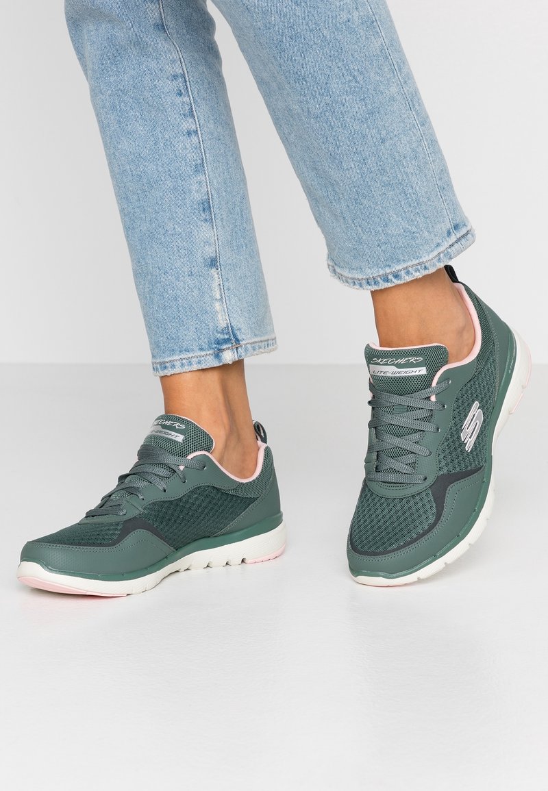 Zelené síťované tenisky s gumovou podrážkou, vybavené růžovými akcenty, zaoblenou  špičkou a logem na straně. Pár je nošený s světle modrými džíny.