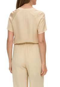 Blouse à manches courtes en tissu beige doux, avec une coupe décontractée et des plis discrets dans le dos, associée à un pantalon beige légèrement froissé.