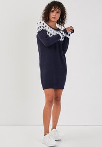 Robe pull marine avec détail de yoke imprimé blanc, manches longues et poignets côtelés. Texture douce, design à longueur genou, associée à des baskets blanches.