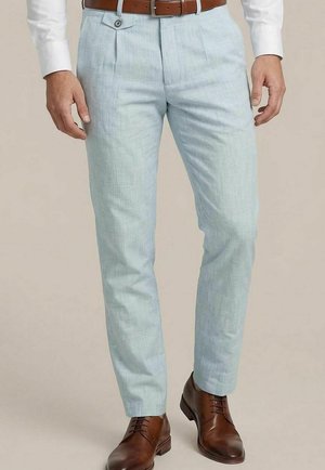 Man draagt lichtblauwe nette broek, bruine leren riem, wit overhemd en gepolijste bruine leren nette schoenen, staand tegen een neutrale achtergrond.