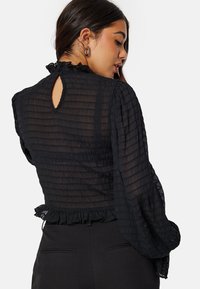 Blusa nera textured con maniche lunghe, scollatura arricciata e dettaglio a chiave sulla schiena. Il tessuto ha un aspetto stropicciato.