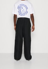 Bílé tričko s modrým grafickým textem a ilustrací na zádech; černé cargo kalhoty s elastickým pasem a bočními kapsami.