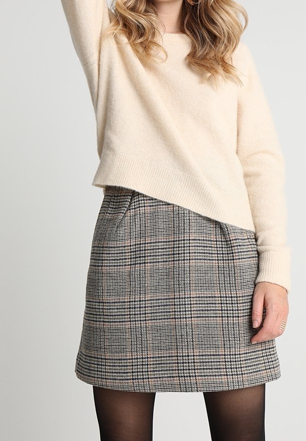 Femme portant un pull crème et une mini-jupe à carreaux grise avec des collants noirs, posant avec un bras levé sur un fond clair.