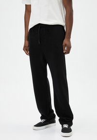 Pantalones deportivos negros con cintura ajustable, piernas ajustadas y textura suave, combinados con zapatillas negras y una camiseta blanca.