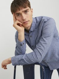Camisa de botones azul con tejido texturizado, de mangas largas y botones oscuros. El modelo se apoya en una silla azul, mostrando una pose relajada.
