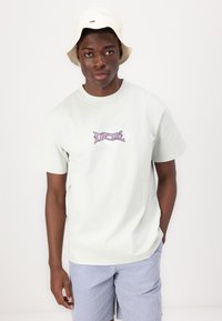 Camiseta de algodón verde claro con cuello redondo y mangas cortas, que presenta un logo púrpura de "Rip Curl" en el pecho. Combinada con pantalones cortos a rayas.
