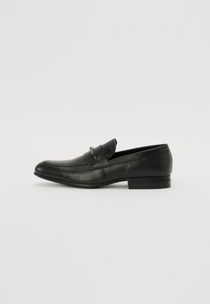 Sorte skinn loafers med avrundet tå, slip-on design, liten metallisk detalj på fronten, og lav, stablet hæl.
