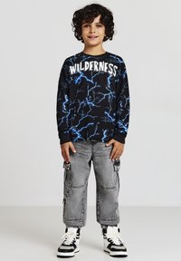Maglietta nera a maniche lunghe con grafiche di fulmini blu e testo "WILDERNESS", abbinata a pantaloni cargo grigi e sneakers nere e bianche.
