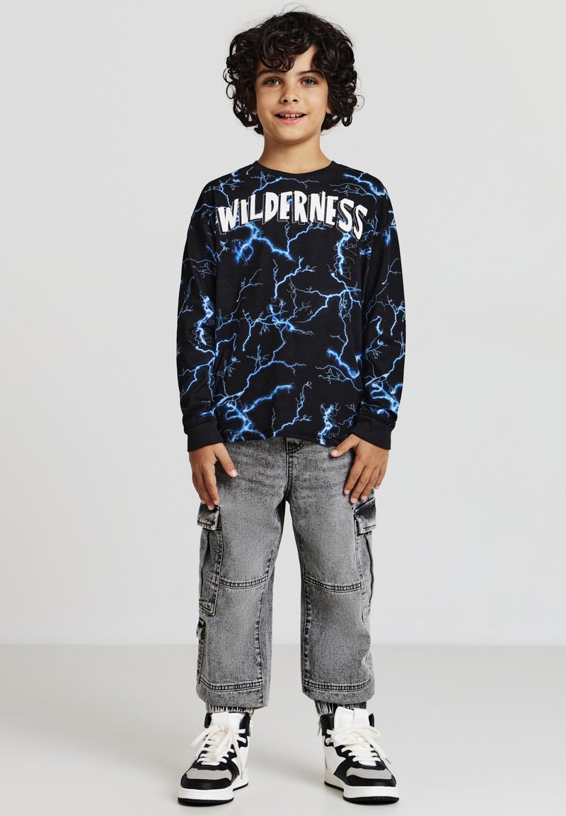 Maglietta nera a maniche lunghe con grafiche di fulmini blu e testo "WILDERNESS", abbinata a pantaloni cargo grigi e sneakers nere e bianche.