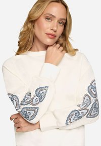 Femme blonde portant un sweat-shirt blanc avec une broderie cachemire bleue et brune sur les deux manches, posant avec une main près du visage.