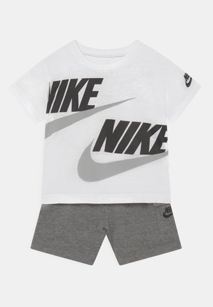 T-shirt bianco in cotone con grandi loghi "NIKE" neri e grigi; abbinato a pantaloni corti grigi con un piccolo logo nero sulla tasca.
