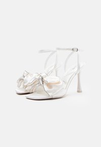 Biele saténové sandále na vysokom opätku s volánikmi a kryštálovými akcentmi, nastaviteľným popruhom na členku a elegantným otvoreným špičkou.