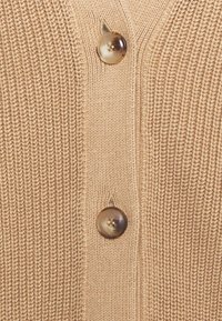 Beige stickad kofta med knappstängning och en glänsande, rund knapp. Texturen är ribbad, vilket förbättrar dess taktila utseende.