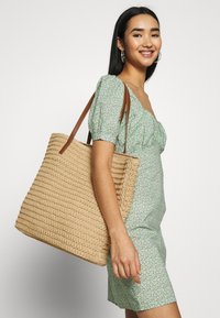 Beige vævet tote taske med brune stropper, der har et tekstureret mønster. Model iført en grøn blomstret kjole med puffærmer og smilende.