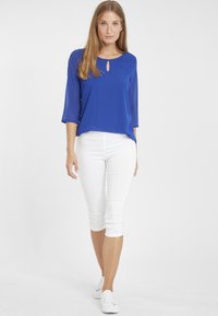 Koninklijk blauwe blouse met driekwartmouwen en een keyhole-detail, gecombineerd met witte aansluitende capri-broeken en witte sneakers. Glad materiaal.