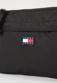 Svart nylonp poses med glidelås, vevd stropp og et rektangulært Tommy Jeans-logo i blått, hvitt og rødt.