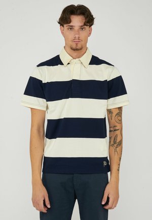 Giovane uomo con capelli mossi che indossa una polo a righe orizzontali blu navy e bianche, in piedi davanti a uno sfondo chiaro e semplice.