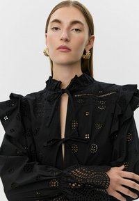 Blusa negra con mangas con volantes, que presenta patrones de encaje con recortes y un lazo en el escote en forma de ojal. Tejido texturizado con detalles intrincados.