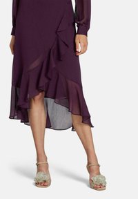 Robe violette avec ourlet évasé et superposition transparente, accompagnée de sandales dorées claires à lanières avec un détail floral sur l'orteil.