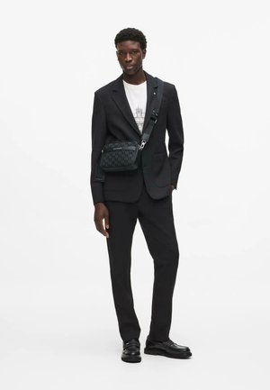 Man in zwart pak, wit grafisch T-shirt, zwarte crossbodytas en zwarte loafers, staand tegen een effen witte achtergrond.
