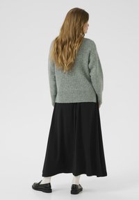 Vrouw met lang bruin haar, gekleed in een grijze gebreide trui, een zwarte lange rok, witte sokken en zwarte instappers, staand met de rug naar ons toegekeerd.
