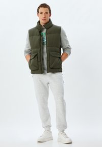 Oliven grøn polstret vest med høj krave, forlommer, båret over en grå sweatshirt og grå sweatpants, parret med hvide sneakers.