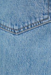 Tela de denim azul claro que muestra un tejido texturizado, con costuras visibles en un color naranja contrastante y un detalle de bolsillo parche.