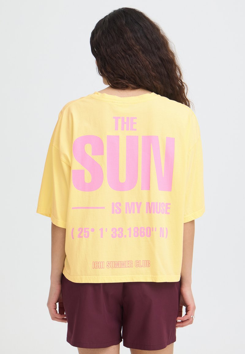 Femme aux longs cheveux bouclés portant un t-shirt jaune ample avec un texte rose « LE SOLEIL EST MA MUSE » et un short bordeaux, debout de dos.